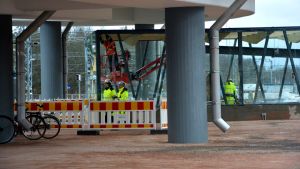 Byggarbetet är ännu i full gång. Nägra byggnadsarbetare arbetar vid en glasinhägnad under en bro.