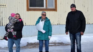 Sanna Kåla, Sirpa Kallio och Jaakko Kallio har alla tre barn i Pettersbacka skola. 2,5-åriga Matilda i mamma Sannas famn är inte riktigt i skolåldern ännu. Tre personer, varav den ena håller ett barn i famnen, står och tittar in i kameran. Gammal träbyggnad i bakgrunden som är beigemålad. Det är vinter och snö.