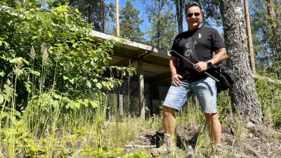 En man i t-skjorta och shorts poserar med en ormkrok i handen framför ett litet hus.