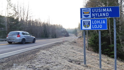 En skylt där det står Nyland och en annan där det står Lojo vid en väg. På vägen kör en bil.