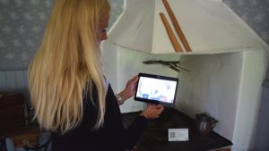Marina Saanila visar hur man rostade kaffe förr i tiden. De gamla redskapen står på vedspisen. Marina Saanila framför en gammaldags vedspis med en ipad i handen.