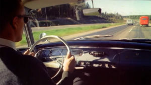 Mies ajaa autoa vuonna 1961 valmistuneessa matkailufilmissä.