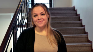 Många väljer att stanna hemma på grund av coronapandemin, men det kommer ändå någon varje dag, berättar Elli-Maija Masanen. Kvinna med mörkblont långt hår och svart kofta och beige t-skjorta står i en trappa och tittar in i kameran. Hon är i 20-årsåldern.