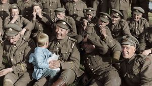 Stillbild från They shall not grow old. Soldater sitter uppställda och spelar munspel och skrattar, en av soldaterna håller ett litet barn i famnen.