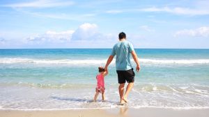 isä ja lapsi kävelevät käsikädessä rannalla vesirajassa