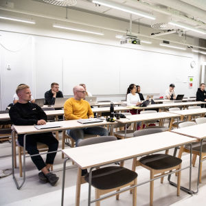 Ett tiotal studerande sitter glest i en föreläsningssal. Till vänster sitter en man i gul tröja, till höger står läraren.