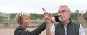 Rabbe Dahlqvist. En kvinna i ljust kort hår har hörlurar på öronen och håller i en mikrofon som är riktad mot en man i mörk väst och rutig skjurta.a och skärmmössa.