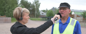 Pauli Koljonen intervjuades i direktsändning av Minna Almark. En kvinna i ljust kort hår har hörlurar på öronen och håller i en mikrofon som är riktad mot en man i gul overall med blå tröja och skärmmössa.