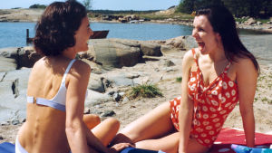 Ihanat naiset rannalla. Kaksi naista istuu rannalla uimapuvuissaan, pyhkeen päällä ja nauraa.