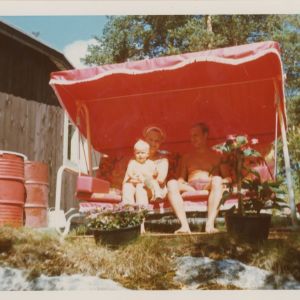 Helena Ahti-Hallberg alle 2-vuotiaana vanhempiensa kanssa aurinkoisella mökkipihalla puutarhakeinussa.