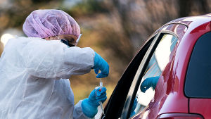 Sjukskötare i hetluften - tar ett prov på en drive-in coronatest. En person i skyddskläder tar ett coronavirusprov med en bomullspinne av en person i en bil med nedvevad bilruta.