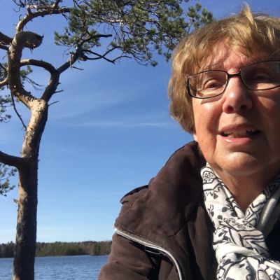 en äldre kvinna som sitter vid havet och tittar mot kameran.