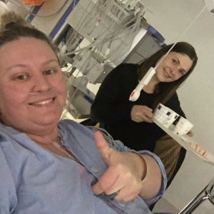 Heidi fick besök av vänner och familj på sjukhuset och det var väldigt viktigt för henne. En kvinna ligger i en säng iklädd sjukhuskläder. Hon visar tummen upp. I bakgrunden syns en annan kvinna som är hennes vän.
