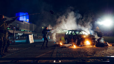 Jasper Pääkkönen står vid en brinnande bil och siktar med ett vapen, inspelningsbild från filmen Omerta 6/12.