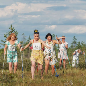 Viisi kukkamekkoihin pukeutunutta henkilöä, heidän joukossaan juontajat Hannamari Hoikkala ja Nicke Aldén, hiihtävät kilpaa suolla.