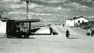 En svartvit bild av en bro och en shell-bensinmack. En kvinna cyklar över bron.