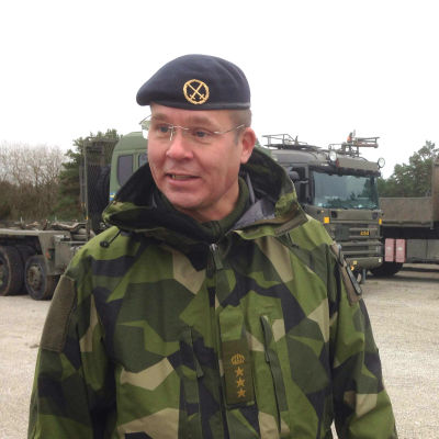 Mattias Ardin, leende, ser förbi kameran, militära lastbilar i bakgrunden.