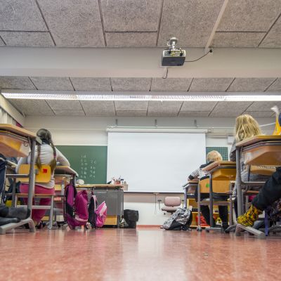 Koululuokka istuu pulpeteissa. Tunnistamattomia lapsia kuvattuna takaa.