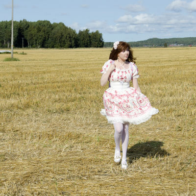 En kvinna klädd i så kallade Lolitakläder - en fluffig vit och rosa klänning med spetsar på. Hon har vita högklackade skor och blommor i håret. Hon går på en nyklippt åker. 