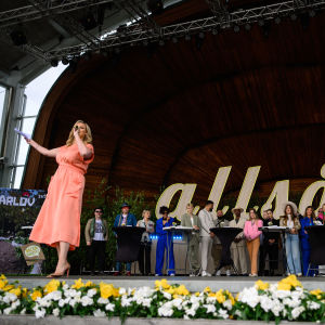 blond dam iklädd klänning står på scen och talar i mikrofon och och presenterar flera andra artister som står bakom henne