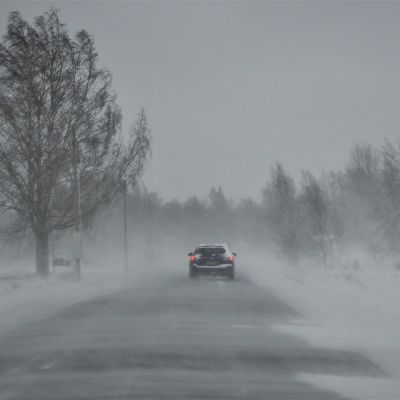 Bil på väg längs landsväg i snöyra och hård vind.