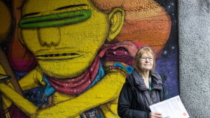 Marjatta Uusitalo står framför en muralmålning längs med cykelvägen Banan i Helsingfors. I handen har hon papper med anvisningar för hur man ska gå till väga för att ansöka om tillstånd för en muralmålning i Helsingfors. Anvisningarna hittar man också på Helsingfors webbplats. En kvinna står framför en väggmålning.