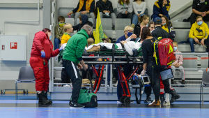 Grankullaspelaren Jonathan Berg lämnar planen på bår. En spelare ligger på bår omringad av flera personer.