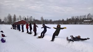 Noin kymmenen henkilöä seisoo lumisessa maisemassa tien reunassa, ja he oat kaatumassa taaksepäin selilleen pehmeään lumihankeen.