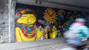 Muralmålningen av Os Gemeos och EGS som finns längs cykelvägen Banan gjordes 2017. En cyklist susar förbi ett gatukonstverk på en vägg.