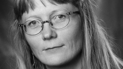 En svartvit bild av en kvinna i medelåldern med långt, blont hår. Hon har glasögon på sig och tittar rakt in i kameran.