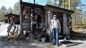 Nu räcker det med korta promenader och att driva ett museum, för Penttis del. Pentti Kronqvist står framför en gammal jaktstuga av trä.