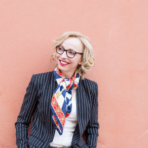 Maria-Elena Ehrnrooth. Maria-Elena Ehrnrooth, en dam med glasögon och ljust, lockigt hår står vid en rosa vägg.