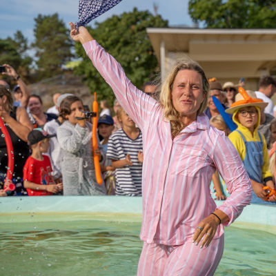 En kvinna i rosa pyjamas står mitt i en vattenfylld fontän. Hon skrattar och vinkar!