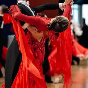 Ett par som dansar rumba. Följaren är iklädd en röd klänning, med håret stramt uppsatt i en knut, och böjer med ansiktet bort från kameran. Ledaren är iklädd kostym.