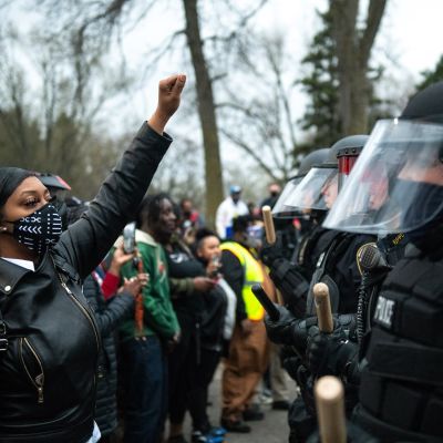 Mielenosoittajien rivistä erottuva tummaihoinen nainen nostaa nyrkkiään ilmaan mellakkavarusteisten poliisien edessä