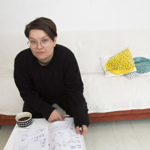 Zarah Holmberg hemma i sin mysiga lägenhet. Zarah Holmberg sitter i en soffa och läser i tidningen hen gjort. Hen tittar in i kameran.