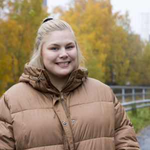 En blond kvinna iklädd en tjock beige dunjacka. Hon ler. I bakgrunden syns träd med gula höstlöv. 