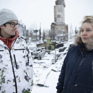 Kuopiolaiset Kimmo ja Nina Perämäki seisovat palaneen omakotitalonsa edessä. Kuvan keskellä palossa pystyyn jäänyt savupiippu.