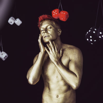 Antti Tuisku kullatussa kropassa ja punaisilla hiuksilla poseeraa mustalla taustalla Kesäkumibiisin videolla. Antin ympärillä on karvanoppia.