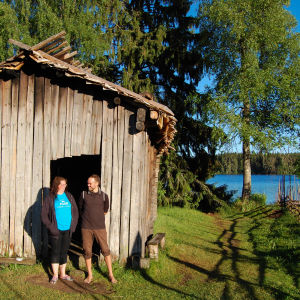 Puinen mallassavusauna järven rannalla, omistajat Merja ja Juha Väinämön seisovat saunan edessä.