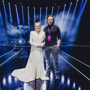 Valkoiseen esiintymispukuun pukeutunut Katri Helena seisomassa sinertävällä UMK-lavalla. Vieressä seisoo vaatesuunnittelija Teemu Muurimäki.
