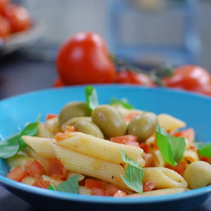 En portion penne pasta med tomatsås, garnerad med oliver och färska örter. Portionen är serverad på ett himmelsblått fat.