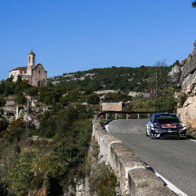 Sebastien Ogier rattar sin rallybil på de slingriga bergsvägarna i Katalonien. I bakgrunden en kyrka på ett berg.