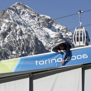 Miehet ripustavat Torinon v. 2006 talviolympialaisten banderollia, taustalla Alppien huippu ja hiihtohissi.