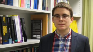 Fredrik Malmberg doktorerade i statsvetenskap förra året. fredrik malmberg står med kavaj vid en bokhylla med statsvetenskaplig litteratur
