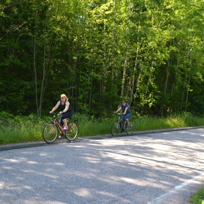Kaarina Hurtta och Sari Siikala cyklar längs med Eriksnäsvägen. I bakgrunden grön skog.