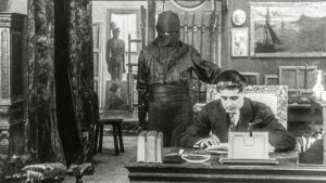 Fantomas. Fantômas seisoo kirjoituspöydän ääressä istuvan miehen takana. Kuva elokuvasarjasta Fantômas