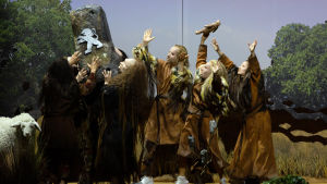 Monista eri ihmislajeista koostuva esihistoriallinen joukko nostaa palvoen päänsä päälle kiven, johon on hakattu Suomen leinoja. Kuvan laidassa kameraa tuijottava täytetty lammas.