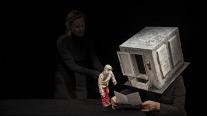Toivo hos arbetsförmedlaren. I bakgrunden animeraren Mila Nirhamo och Iida Vanttaja som arbetsförmedlare. Foto från Kohtauspaikkoja kadonneille på Finlands nationalteater.