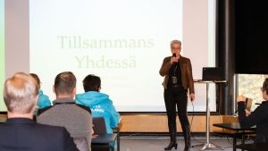Henna Ljung säger att coronaverkligheten blivit det nya normala. En kvinna med kort ljust hår talar inför publik. I bakgrunden en skärm där det står "Tillsammans".